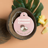 Pink car charm tag with "Bronzed Coconut" text and coconut illustration, displayed in halved coconut shell with tropical leav