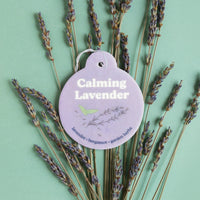Lavender-colored car charm with text and floral design, displayed among dried lavender sprigs on mint green background.
