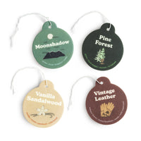 Four circular car air freshener charms in green, black, tan, and brown with scent names and botanical illustrations, attached