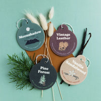 Four round scent car charms labeled Moonshadow, Vintage Leather, Pine Forest, and Vanilla Sandalwood with dried botanicals.