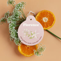 Pink pear-shaped car charm with white floral design, surrounded by dried flowers and orange slices.