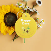 Yellow circular car charm with "Happy Days" text and sunflower design, displayed with fresh sunflowers and daisies.