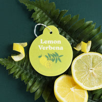 Yellow round car charm with "Lemon Verbena" text and leaf design, white string attachment, displayed with lemon slices and gr