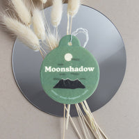 Green circular car charm with "Moonshadow" text, moon and mountain design, attached to white string with dried grass, display