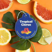 Blue circular car charm with white text reading "Tropical Citrus" and scent notes, surrounded by citrus slices and green leav