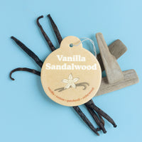 Four car charms with cream-colored round tags, dark vanilla pods, and gray wooden pieces arranged on blue background.