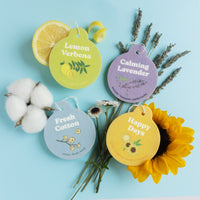 Four round car charms labeled Lemon Verbena, Calming Lavender, Fresh Cotton, and Happy Days, displayed with lemon slices, cot