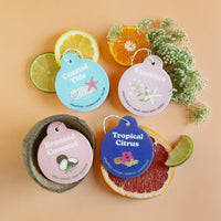 Four round scented car charms labeled Coastal Tide, Florence, Bronzed Coconut, and Tropical Citrus arranged with citrus slice