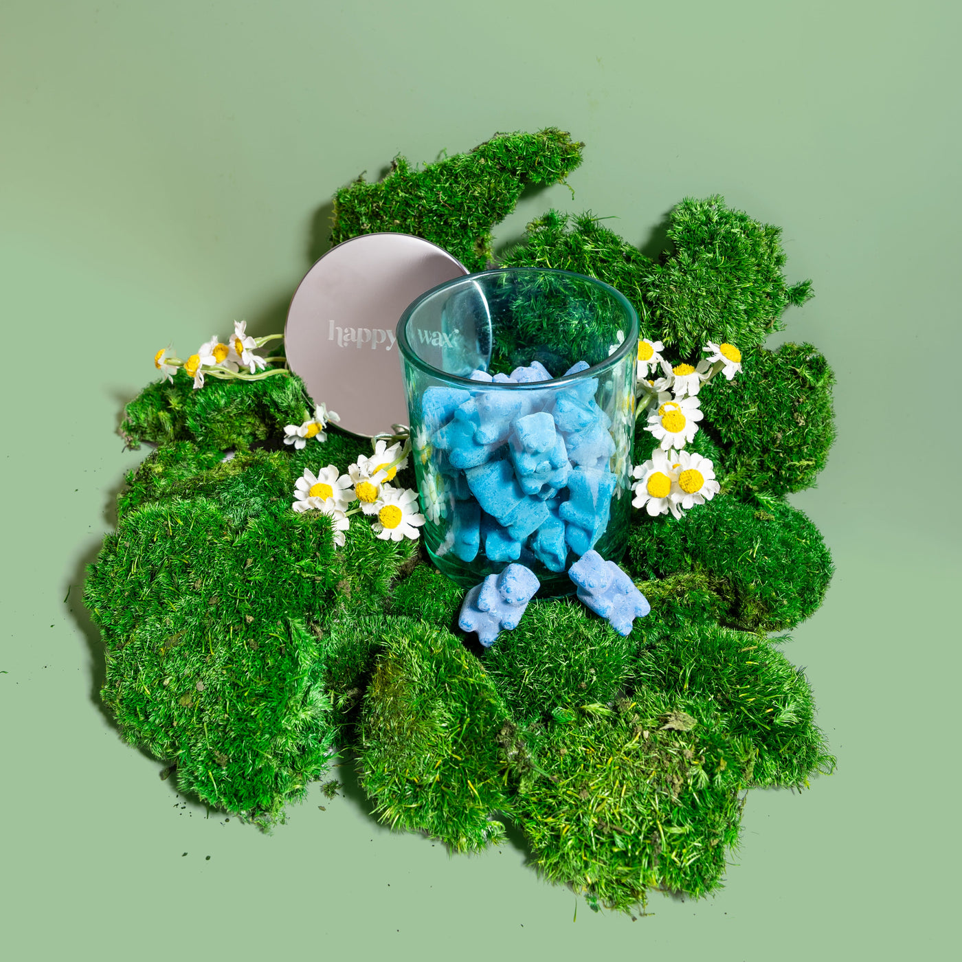 Glass container with blue wax melts on a bed of green moss and white flowers against a light green background.