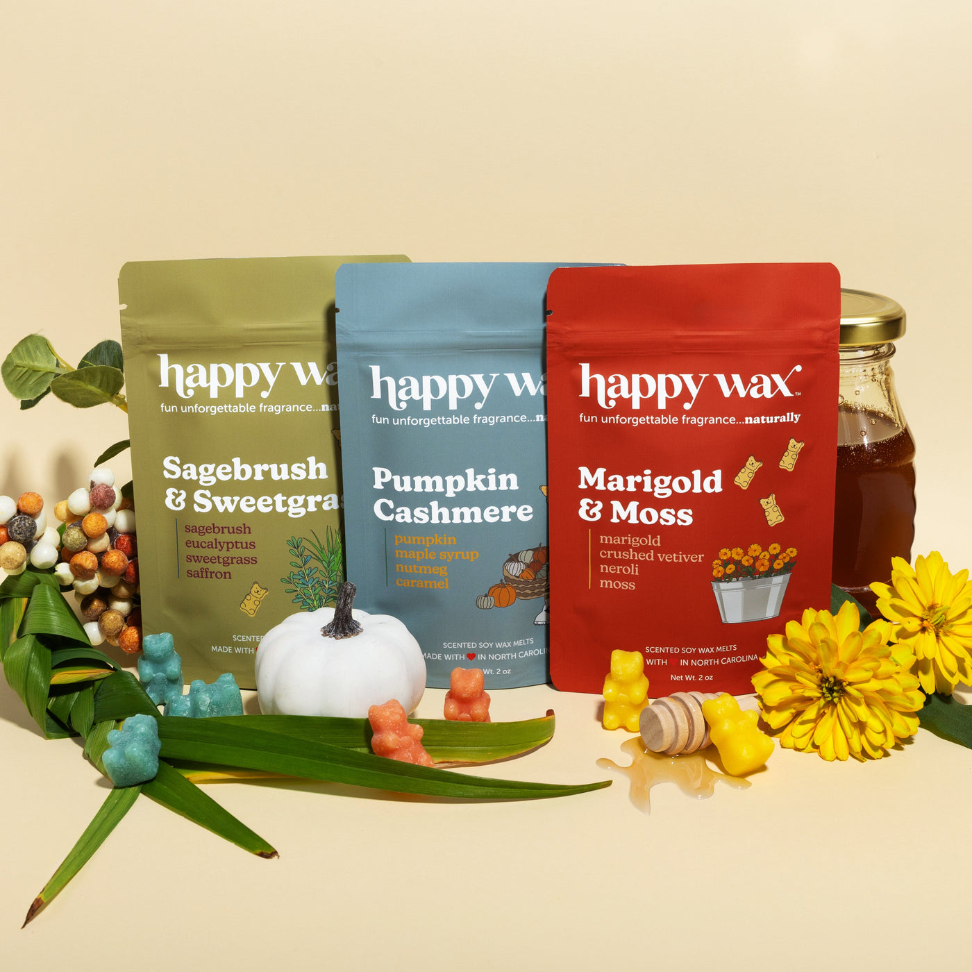 Happy Wax Field Notes 3 pack, Sagebrush & Sweetgrass, Pumpkin Cashmere, and Marigold & Moss, with greenery, yellow marigold flowers, honey, and pumpkin on beige background