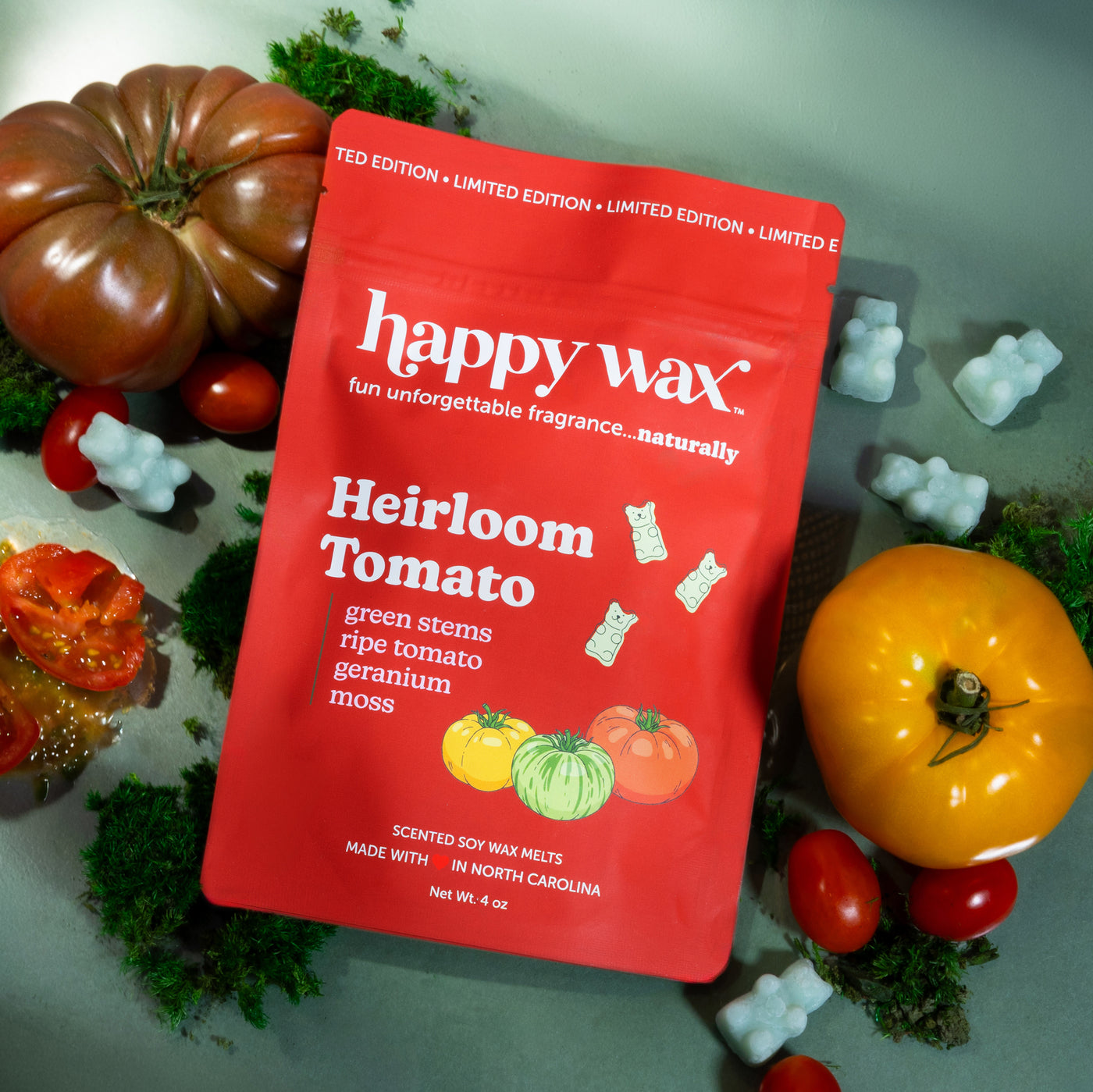 happy wax Heirloom Tomato wax melt packaging surrounded by decorative tomatoes and plants on a green background