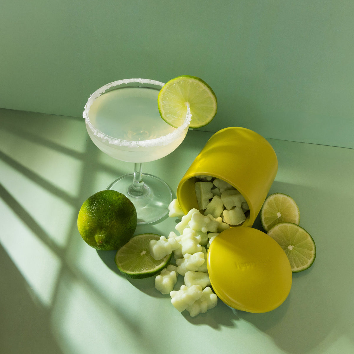 Cocktail with lime slices, wax melts, and a yellow container on a light surface.