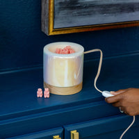 White cylindrical wax warmer with wooden base band, filled with melted wax and pink wax cubes, with power cord.