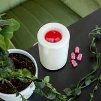 White cylindrical wax warmer with red wax and pink wax cubes on dark surface surrounded by potted plants.