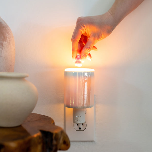 Hand hovering over a cylindrical wall-plug wax warmer with glowing amber light and timer controls on the outlet plate.