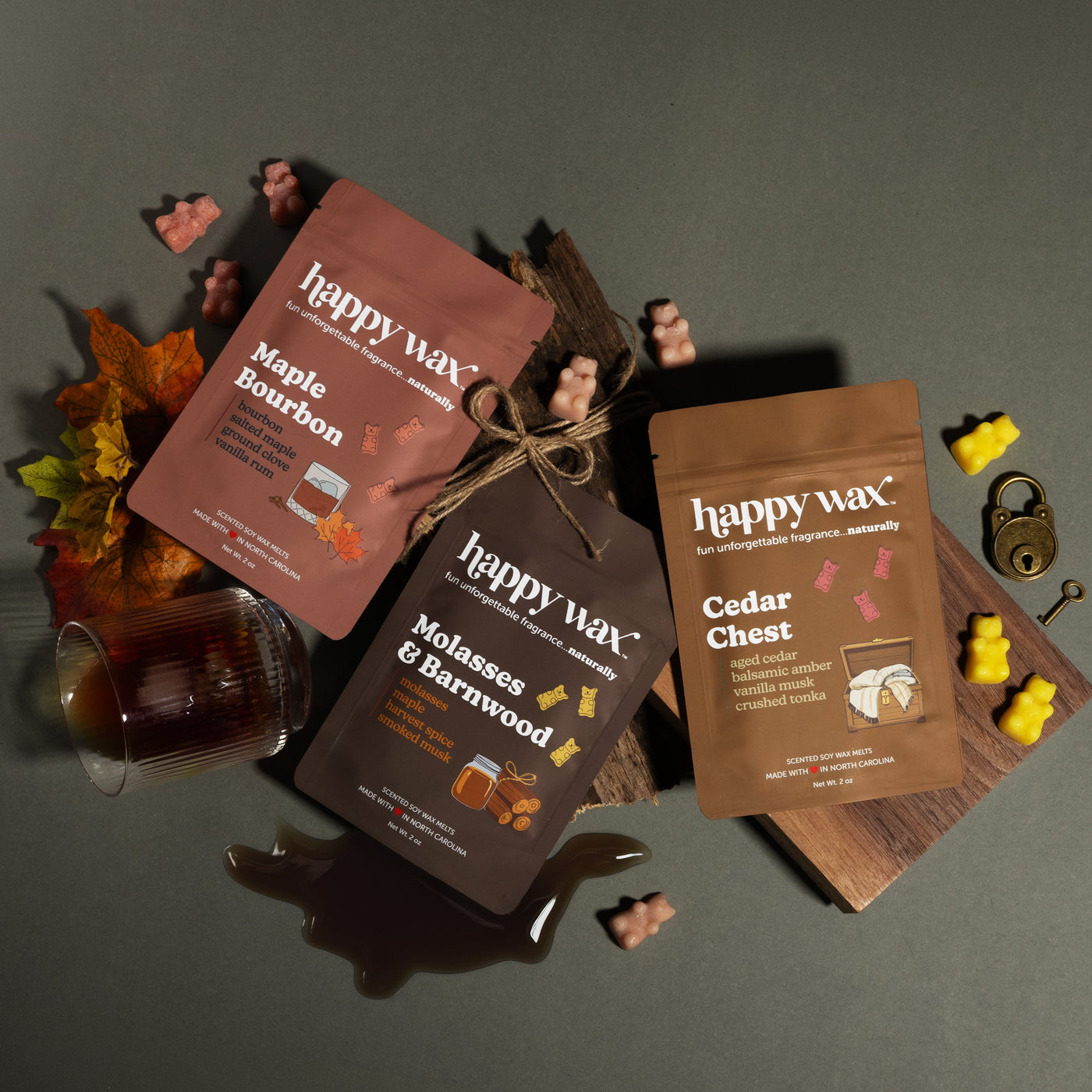 Happy Wax Old Growth 3 pack, Maple Bourbon, Molasses & Barnwood and Cedar Chest pouches with fall leaves, bourbon glass, molasses, and cedar wood on a muted green background