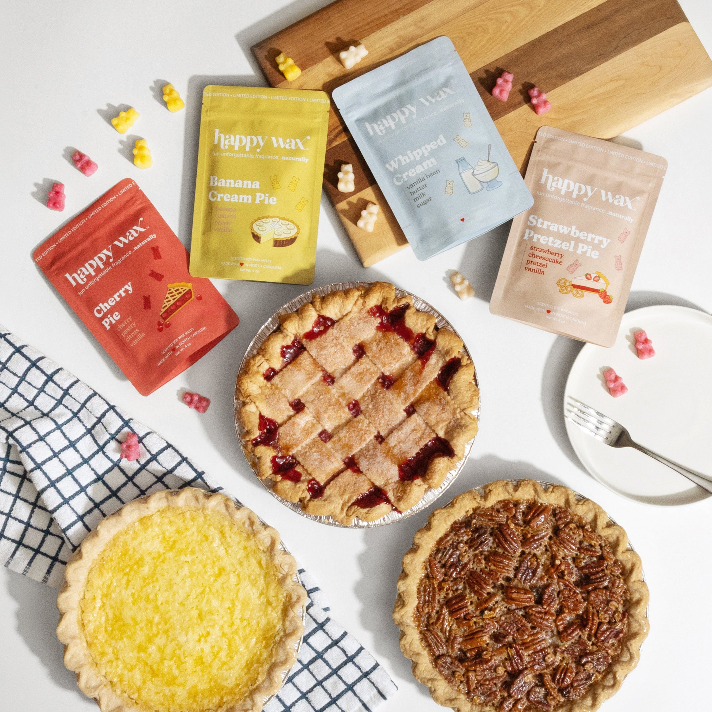 Assorted pies and Happy Wax scented wax melts on a white surface with a wooden cutting board.