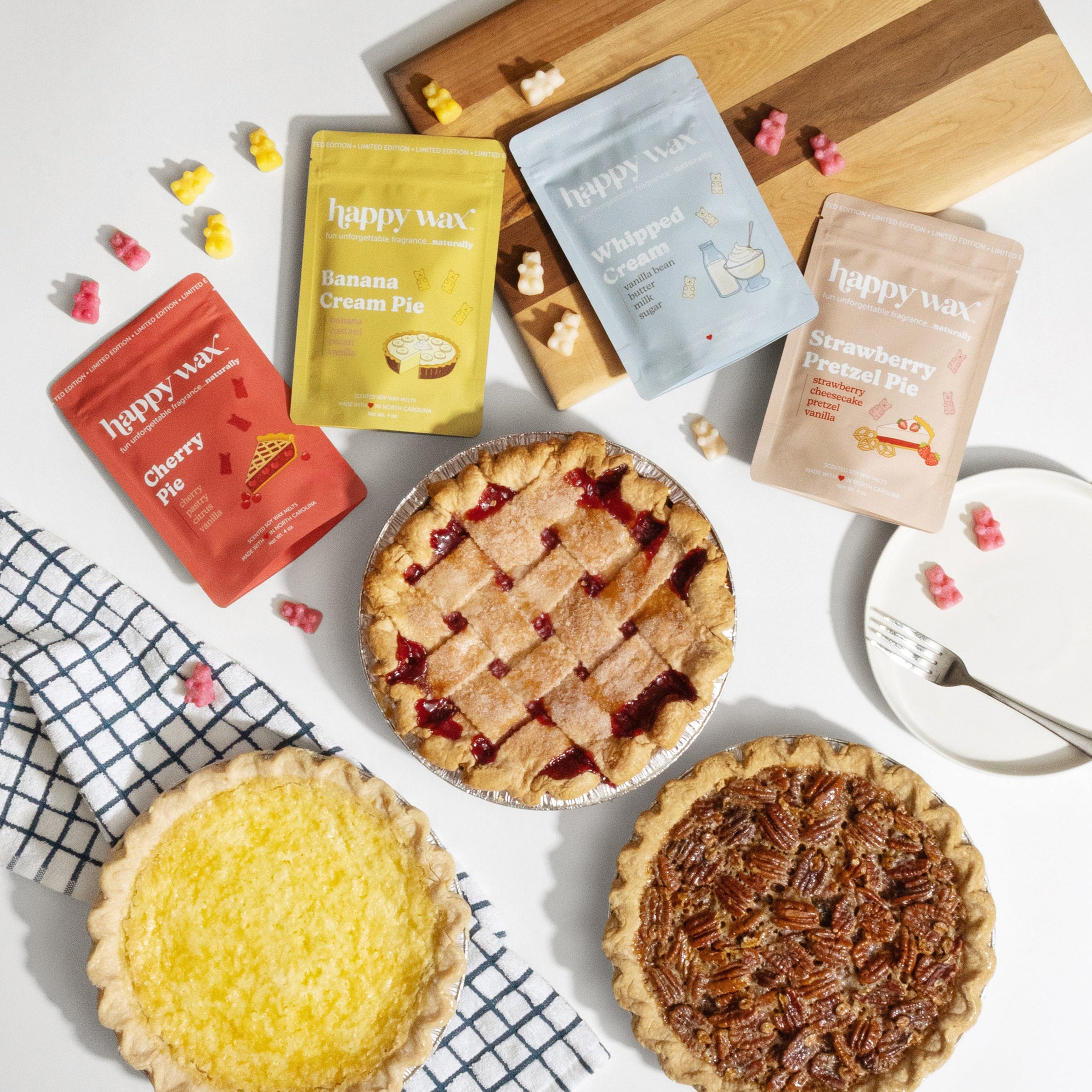 Assorted pies and Happy Wax scented wax melts on a white surface with a wooden cutting board.