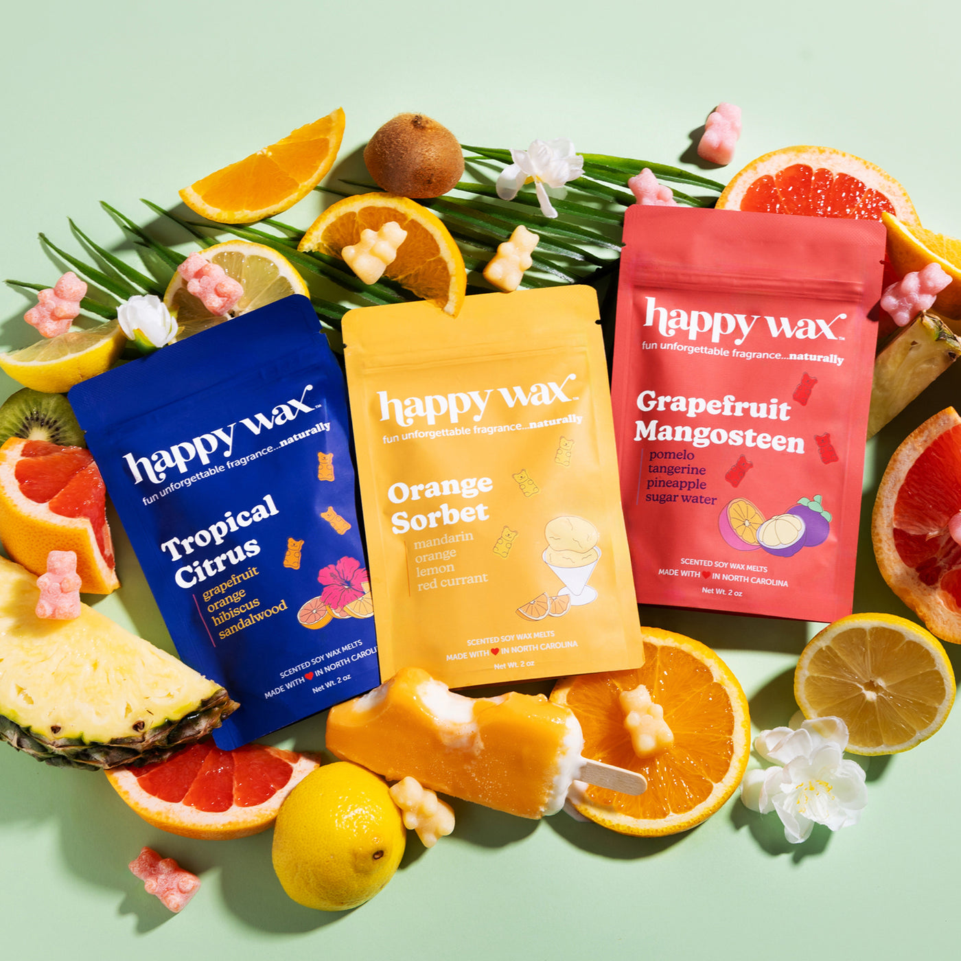 Happy Wax scented wax melts, Tropical Citrus, Orange Sorbet, and Grapefruit Mangosteen surrounded by tropical fruit on light green background.