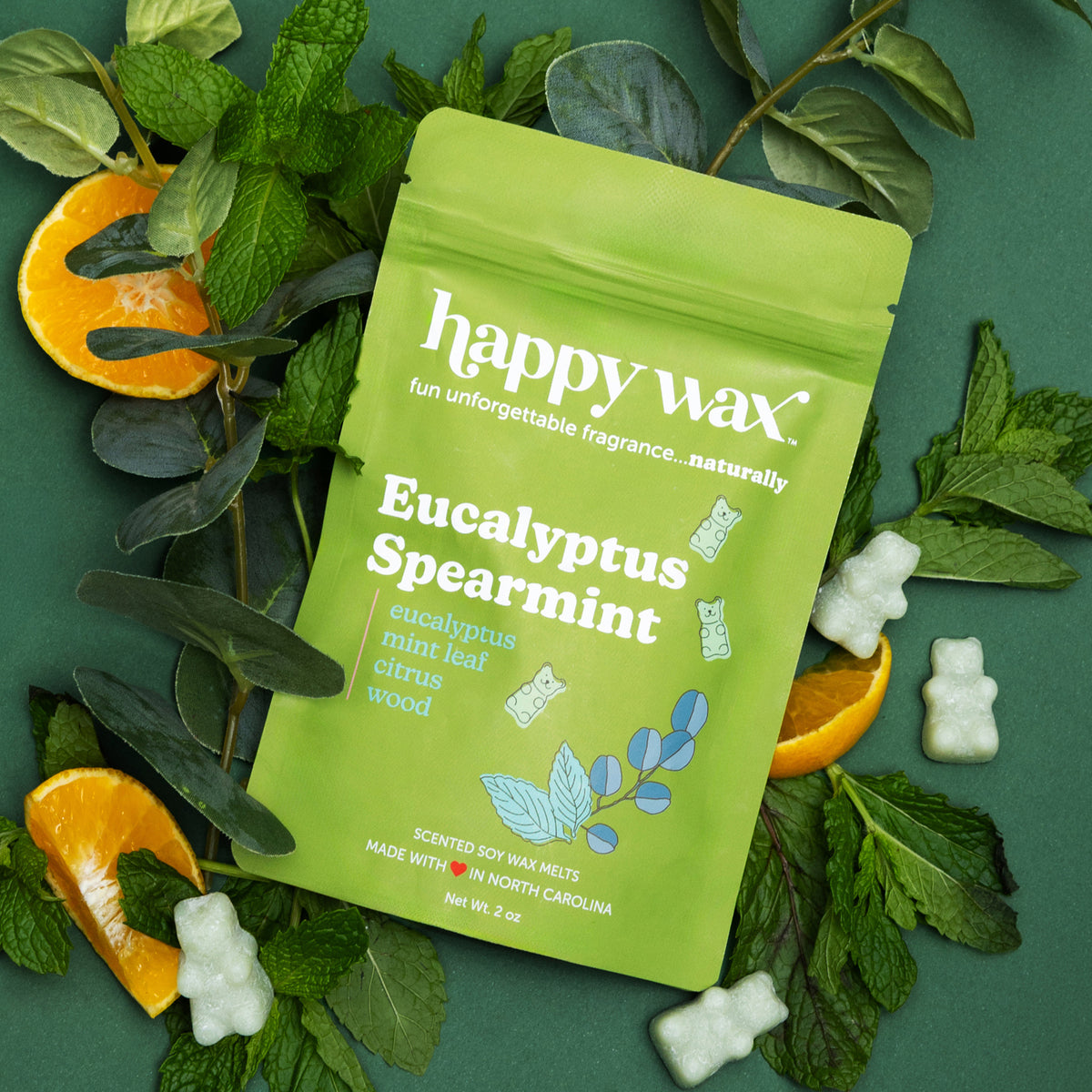 Happy wax scented wax melts, Eucalyptus Spearmint surrounded by eucaluptus, mint leaves, and citrus on a dark green background.