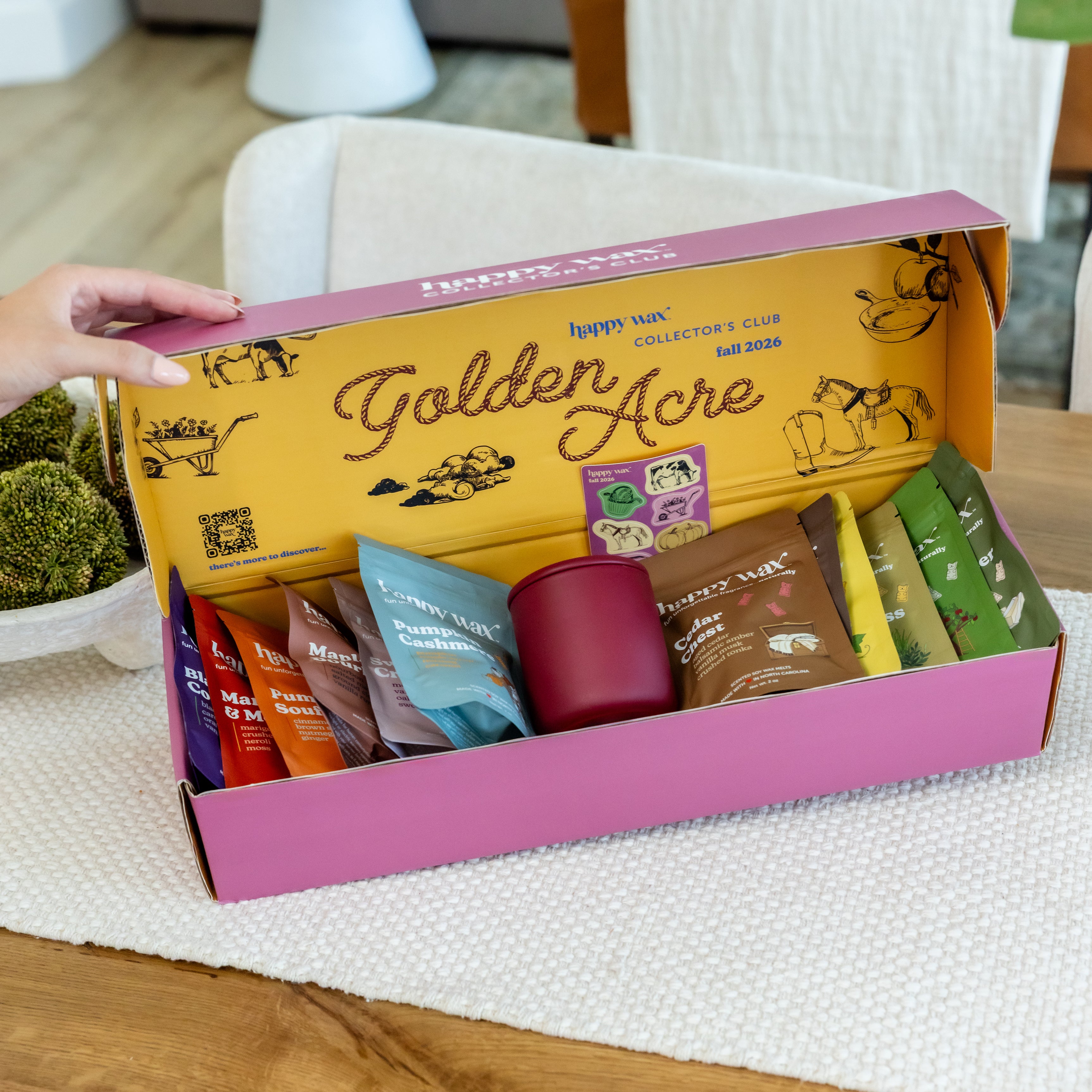Open fall subscription box with yellow lid and pink base containing various products including a burgundy candle, tea packets