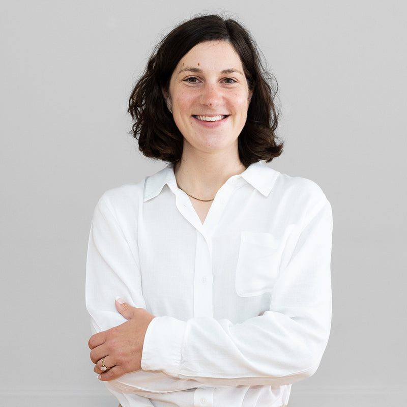 A woman with dark shoulder-length hair stands with arms crossed, wearing a white long-sleeve button-up shirt and beige drawstring pants against a plain light gray wall and wood floor.