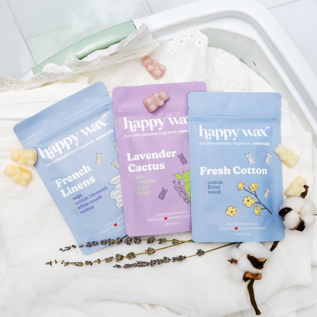 Happy Wax French Linens, Lavender Cactus, and Fresh Cotton pouches in a laundry basket with white linens, lavender, and cotton on white tile floor