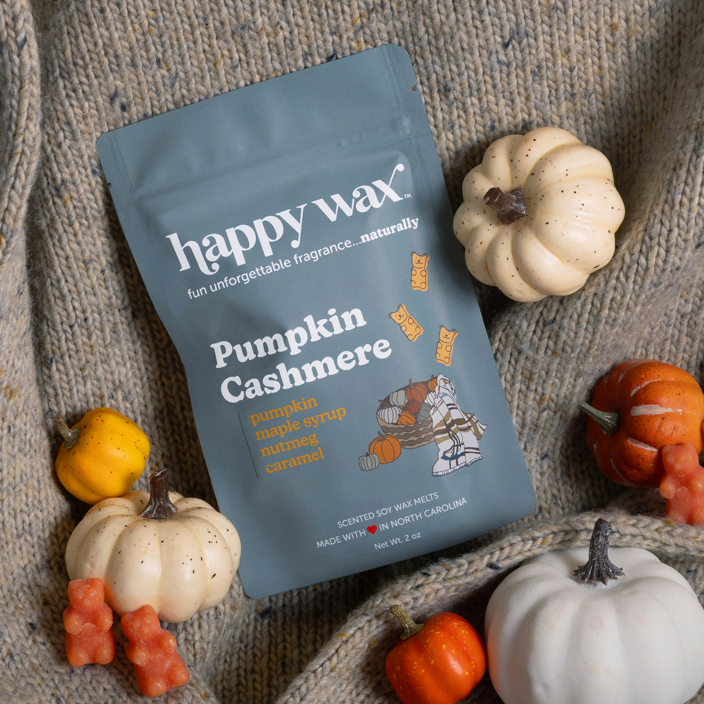 Blue pouch of Pumpkin Cashmere wax melts surrounded by decorative cream and orange pumpkins on a knit fabric background.