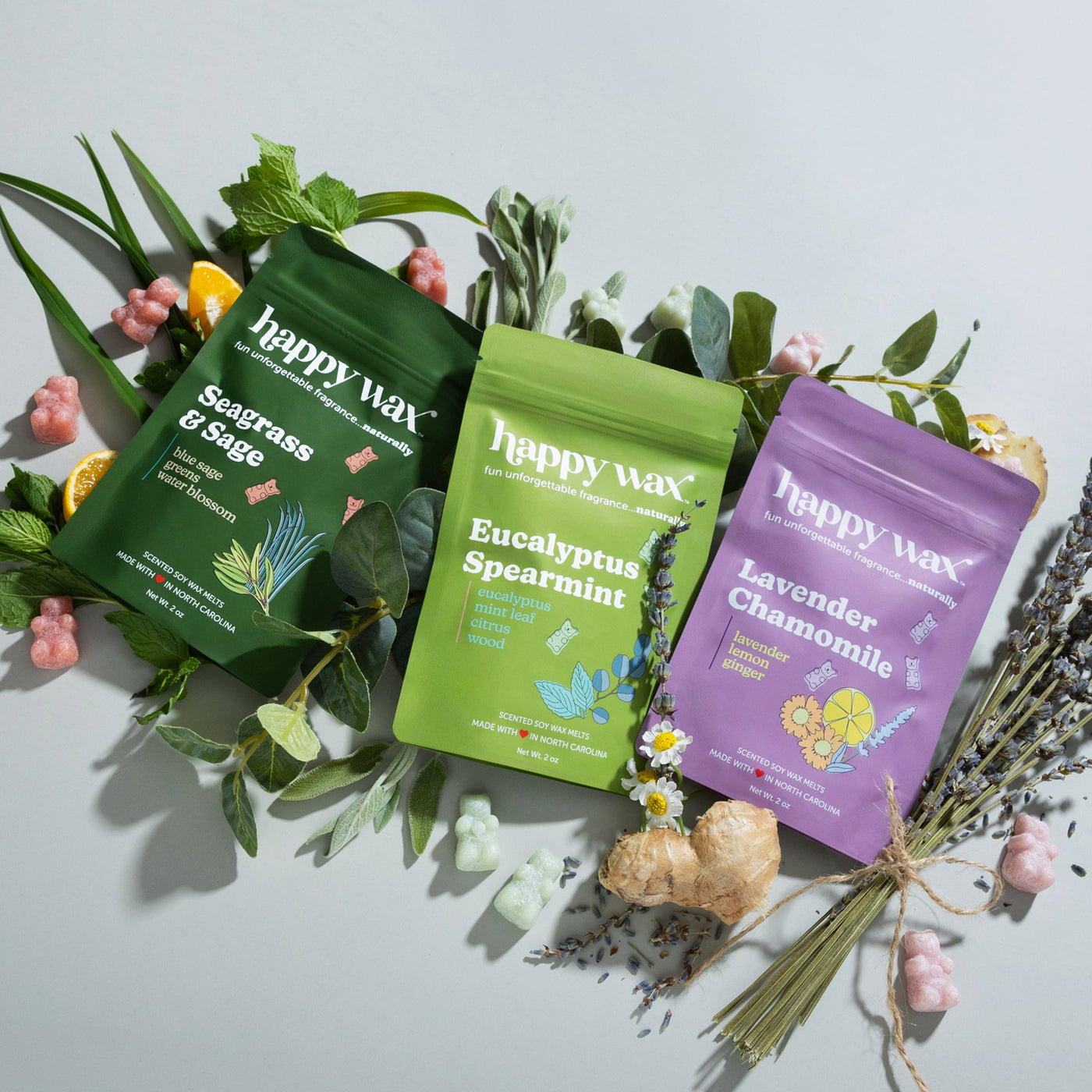 Happy wax scented wax melts, Seagrass & Sage, Eucalyptus Spearmint, and Laventder Camomile surrounded by ingredients - lavender, sage, mint, citrus, ginger, on a grey background.